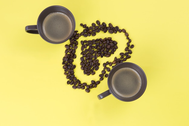 Top view of coffee beans forming heart shapes and cups of coffee isolated on a yellow background