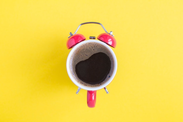 Top view of coffee on the dial of the red alarm clock in the center of the yellow background