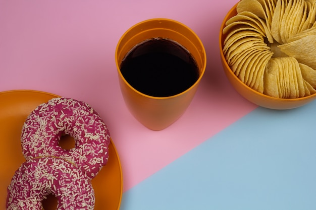 Top view cola glass, chips bowl, doughnut plate , pink and blue background. unhealthy food concept