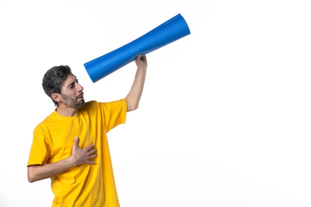 Top view of concentrated young male in yellow shirt holding folded blue mat on white surface