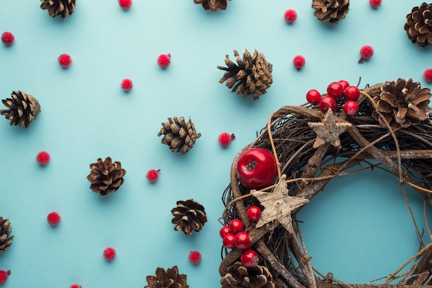 Top view cropped photo of christmas twig wreath scattered holly berries and pine cones on isolated pastel blue background