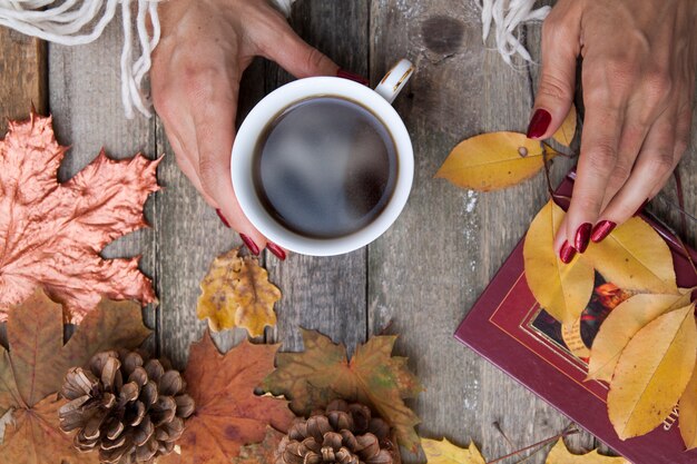 Photo top view cup of coffee in female hands on a wooden table background with autumn leaves, cones