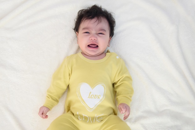 Top view of a cute little Asian baby girl wearing yellow dress, lying on the bed, sad and crying