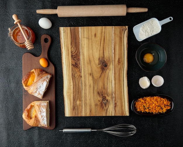 Top view of cutting board with cut croissant quince jam egg orange zest egg yolk egg shell rolling pin on black background