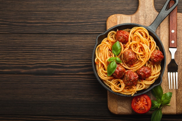 Top view of delicious pasta with meatballs, tomato sauce and fresh basil in cast iron rustic vintage pan served on cutting board, wooden background. Tasty homemade meatballs spaghetti, space for text