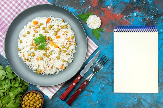 Top view of delicious rice meal with pisum carrot green bundle on purple towel and cutlery set notebook on blue background