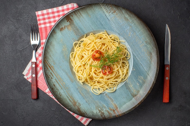 Top view of delicious spagetti served with tomatoes greens and cutlery set on red stripped towel on black background