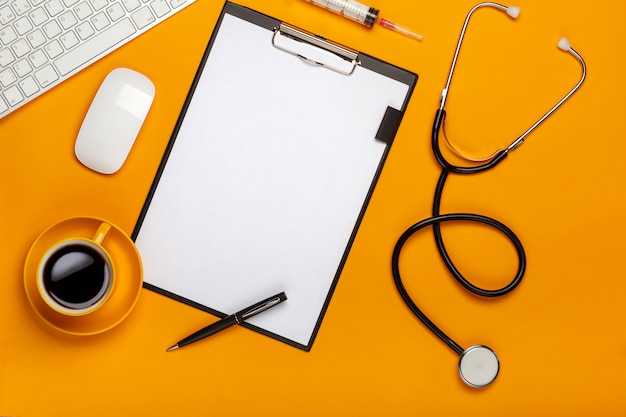 Top view of a doctor's table with notepad and pen stethoscope, keyboard, prescription and pills, a cup of coffee on a yellow background