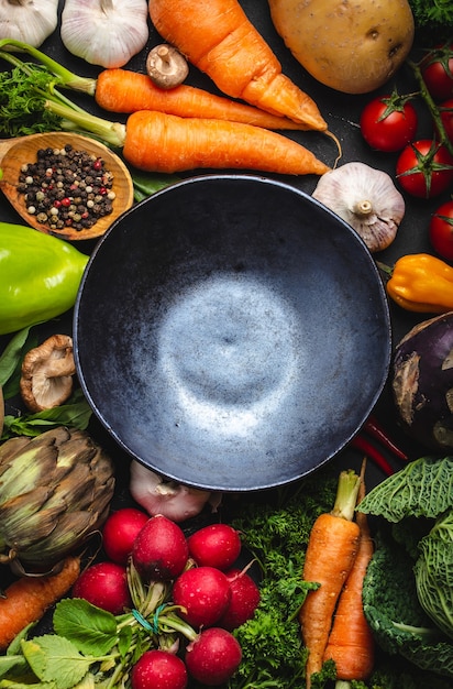 Top view of empty rustic ceramic bowl and fresh farm organic vegetables on rustic black concrete background. Autumn harvest, vegetarian food or clean healthy eating concept with space for text