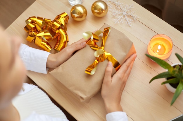 Top view of faceless female holding in hands christmas gift with gold bow on background of table with christmas ribbons and balls preparing for New Year holidays Merry Christmas