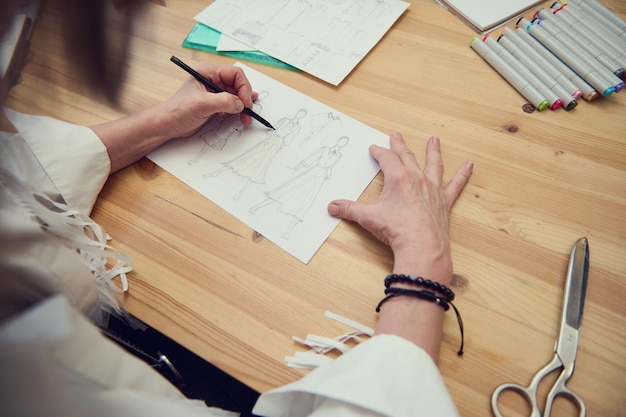 Top view of a fashion designer drawing sketch of new clothes collection in design studio woman tailor creating line working with pencil and watercolor markers on wooden personal desk