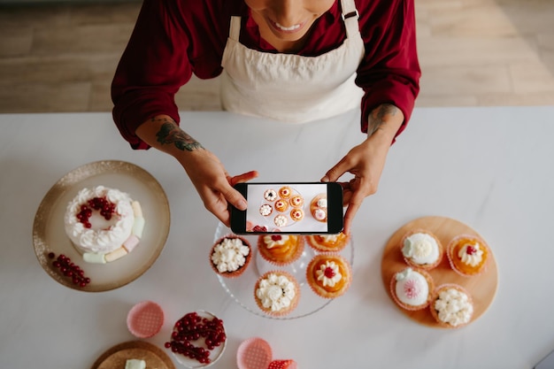 Photo top view of female baker using smart phone while photographing sweet food laying on the table