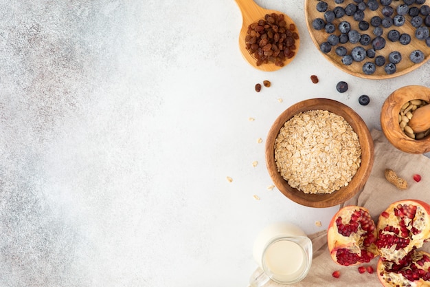 Top view and flat lay of oatmeal porridge in wooden ware with milk, red currant, blueberries, almonds, and garnet on textured concrete light table. 