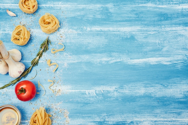 Top view, flat lay. Pasta nests, oil, salt, pepper, tomato, garlic and fork on a blue wooden table.