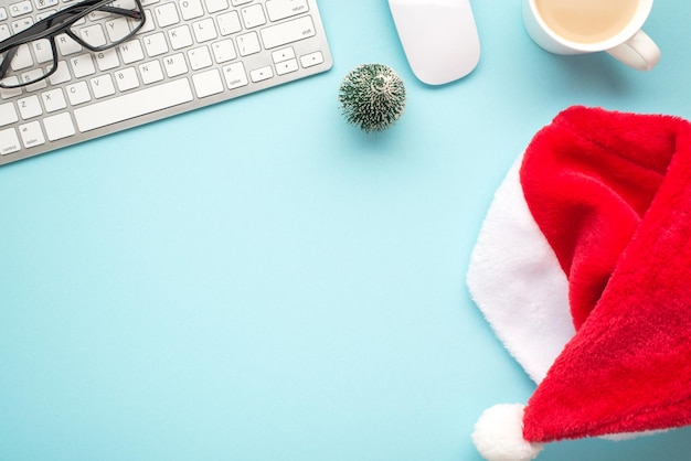 Top view flat lay photo of santa claus hat keyboard computer mouse glasses pine toy and cup of hot drinking on isolated pastel blue background with empty space