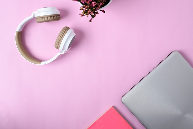 Top View Flatlay of modern wireless headphone, minimalist laptop, Cute purple Flower, and book on pink color workspace