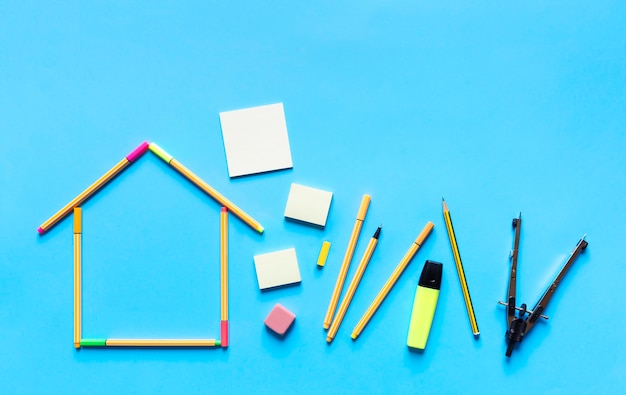 Top view of fluorescent marker pens forming a drawing of a house and other stationery materials on pastel blue background.