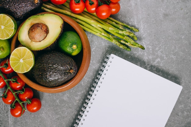 Top view fresh healthy vegetables in wooden plate and blank notebook on gray 