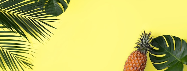 Top view of fresh pineapple with tropical palm and monstera leaves on yellow table background.