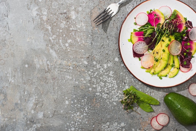 Top view of fresh radish salad with greens and avocado near fork and vegetables on grey concrete