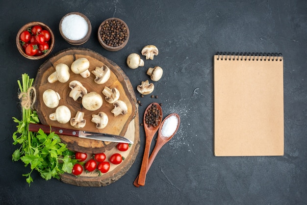 Top view of fresh raw mushrooms and green bundle knife tomatoes spices spiral notebook on wooden board towel on black background