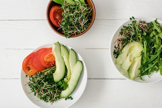 Top view of fresh vegetables with avocado and microgreen in bowls on white wooden surface