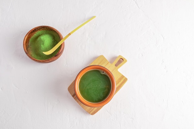 Top view of freshly made matcha green tea in a ceramic cup and matcha powder in a wooden bowl with a bamboo measuring spoon white background