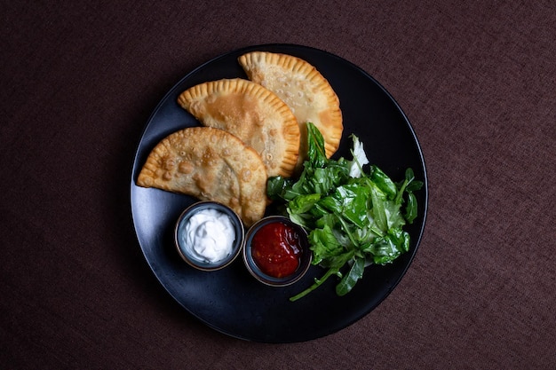 Top view fried chebureks with sauce and herbs on a black plate