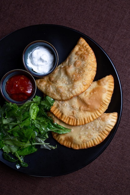 Top view fried chebureks with sauce and herbs on a black plate