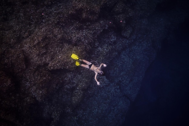 Top view full body of anonymous man in snorkeling mask and yellow flippers swimming under deep dark seawater near massive rocky formation