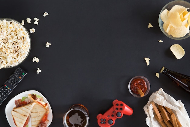 Top view of gamer accessorises and snacks frame, flat lay on black background with copyspace. Joystick and gamepad, keyboard, game console, mouse, mobile phone, beer, chips and popcorn.