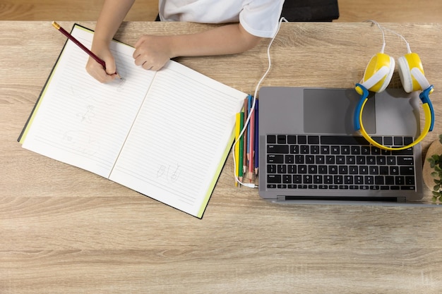 Photo top view of girl kid with headphone and writing on paper book is learning online from  laptop