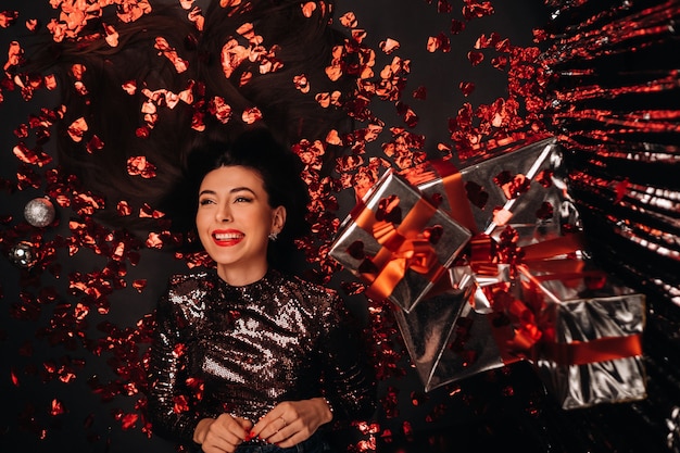 Top view of a girl lying in shiny clothes on the floor in confetti in the form of hearts and gifts.