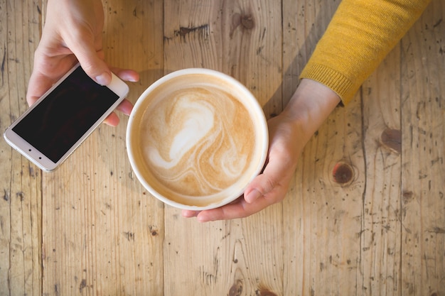 Top view of a girl working on the phone and drinking coffee while sitting at a wooden table