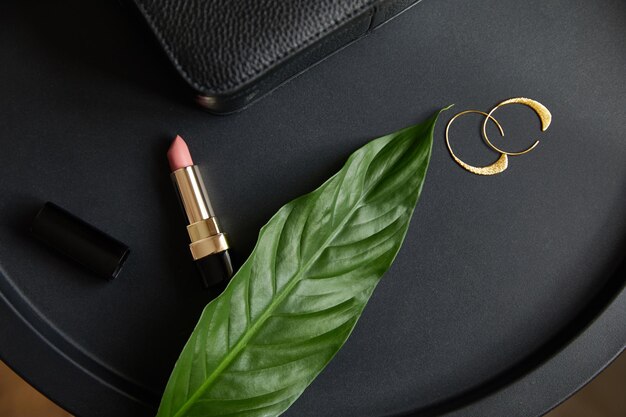 Top view of golden earrings and pink lipstick on black table with tropical leaf