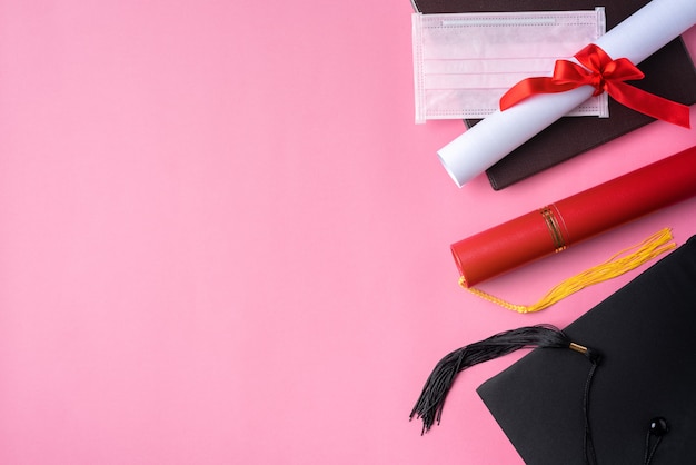 Top view of graduation square academic cap with degree diploma and mask isolated on pink table background.