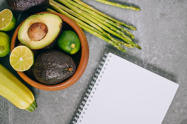 Top view green fresh healthy vegetables in wooden plate and blank notebook on gray 