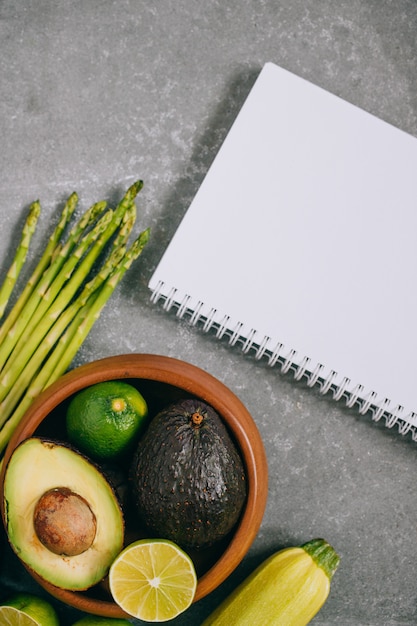 Top view green fresh healthy vegetables in wooden plate and blank notebook on gray 