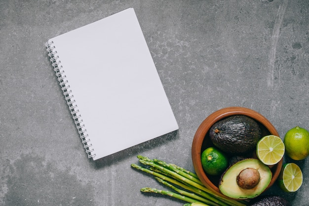 Top view green fresh healthy vegetables in wooden plate and blank notebook on gray