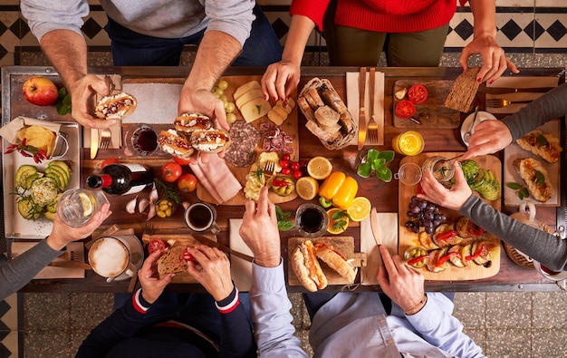 Top view group of anonymous friends eating appetizing various snacks on cutting boards while sitting at wooden table in kitchen