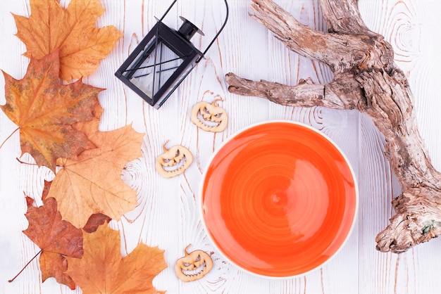 Top view of halloween background with empty orange plate ready to place food lantern colored leaves driftwood on wooden white table and spooky wood carved pumpkins