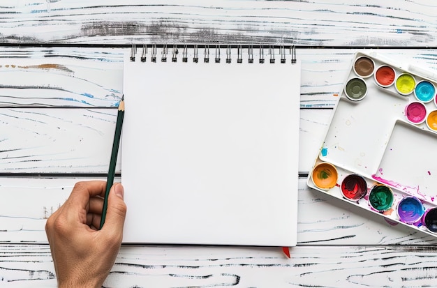 Top view of a hand holding a pencil and sketchbook with watercolor paints on a white wooden tabl