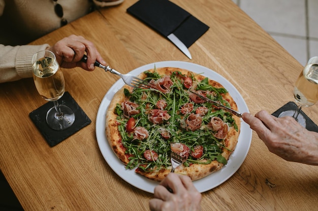 Top view of hands with thin fingers of unrecognisable elderly people possibly women eating pizza and drinking wine or champagne in cafe celebrating their meeting or holiday