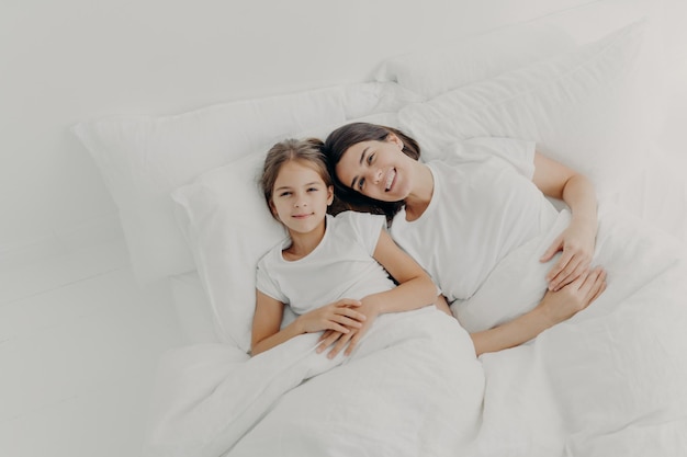 Top view of happy mother and daughter awake in good mood feel relaxed enjoy comfort in bed rejoice good morning dressed in pyjamas lie under white blanket on soft pillows Bedding time