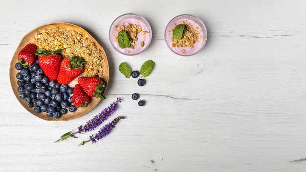 Top view of a healthy breakfast with two bowls of berries yogurt with granola and oats and a wood plate of strawberries and blueberries