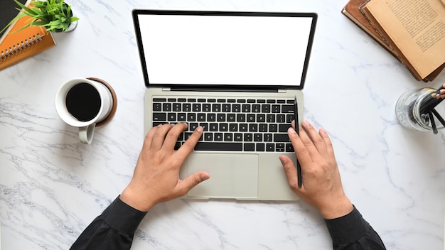 Top view image of businessman's hands typing on computer laptop keyboard with white blank screen that putting on marble texture table surrounded with coffee cup, pencil holder, potted plant and books.