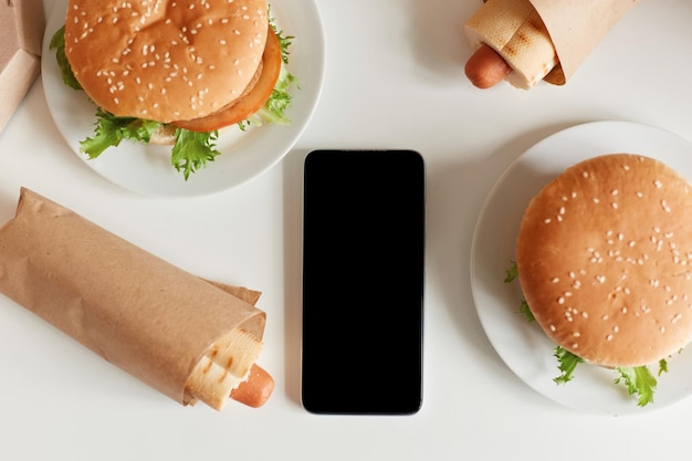 Top view image of junk food on table tasty sausage in dough and big hamburgers smart phone with black blank screen copy space for advertisement or promotional text