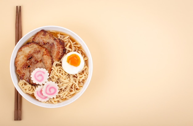 Top view of Japanese noodle soup ramen in white bowl with noodles, meat broth, sliced roasted pork, narutomaki, egg with yolk on pastel beige background. Traditional dish of Japan, space for text