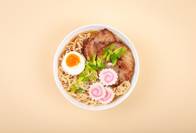 Top view of Japanese noodle soup ramen in white ceramic bowl with noodles, meat broth, sliced roasted pork, green onion, narutomaki, egg yolk, beige background. Traditional dish of Japan, close-up