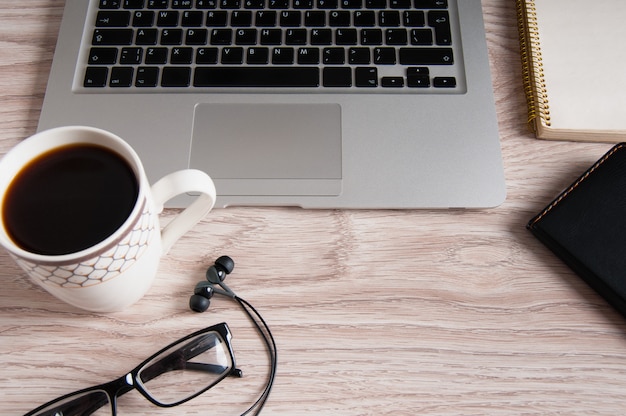 Top view of laptop and notepad, black glasses and headphones on white wood desktop and cup of hot coffee.